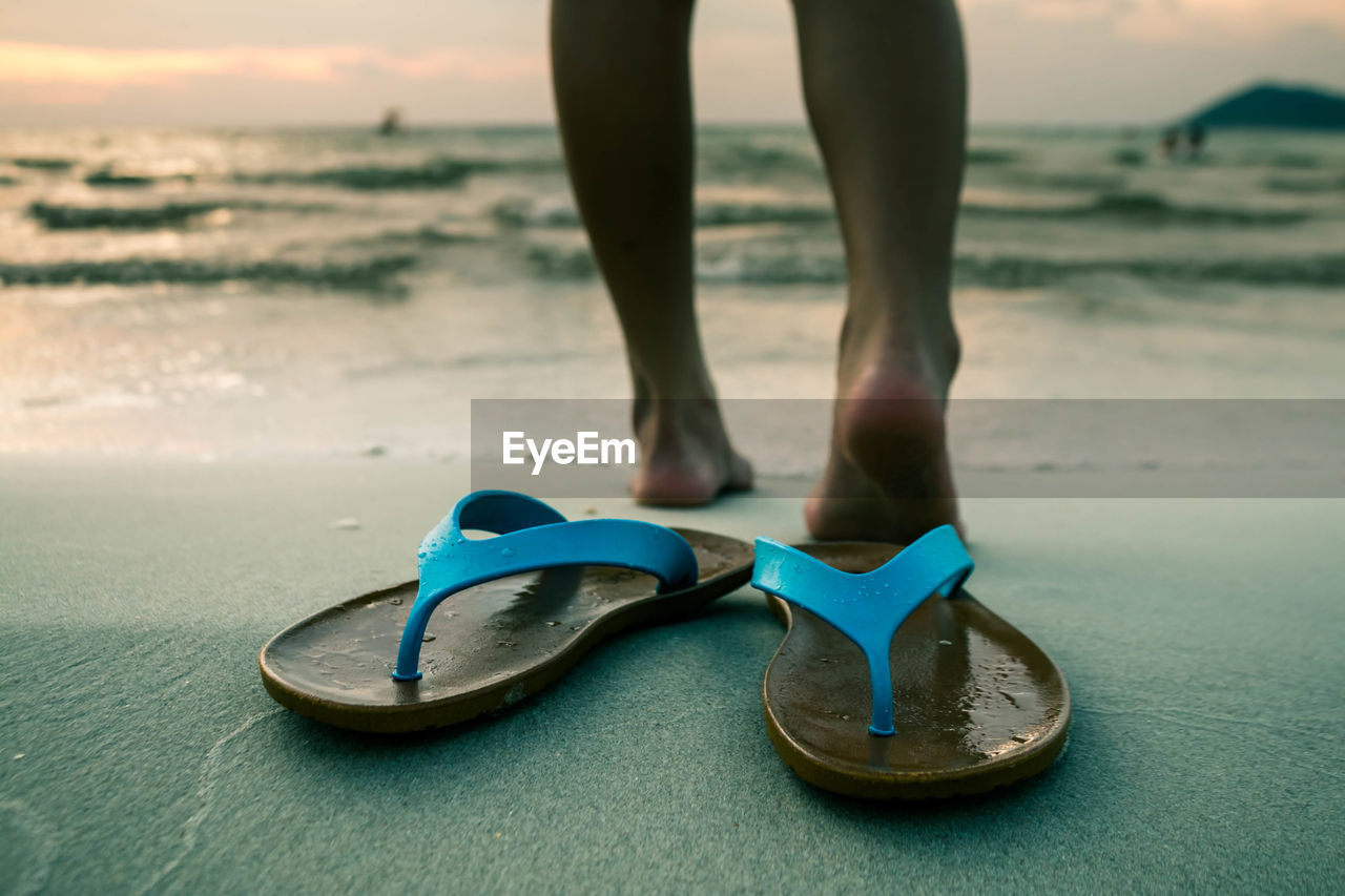 Low section of woman standing on shore with flip-flops in foreground at beach