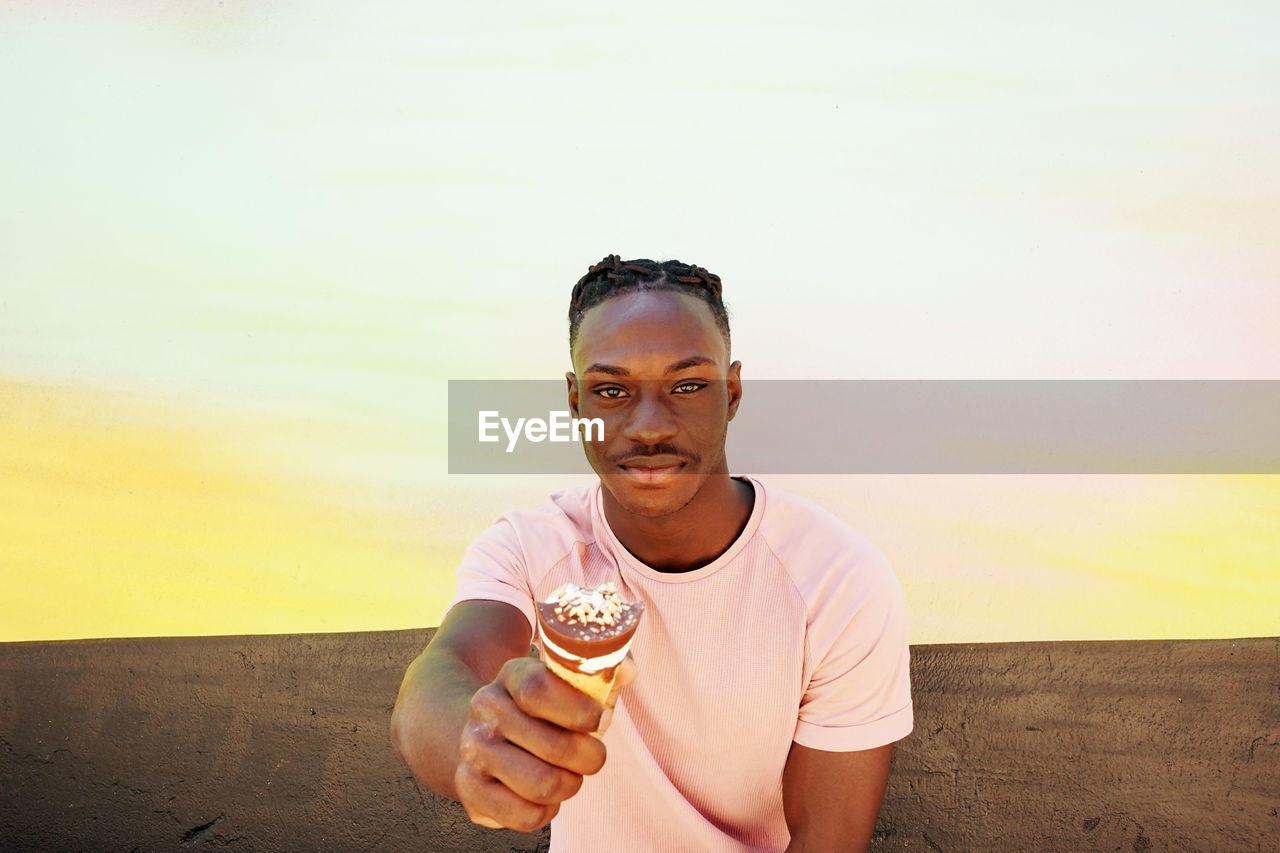 PORTRAIT OF YOUNG MAN HOLDING ICE CREAM AGAINST SKY