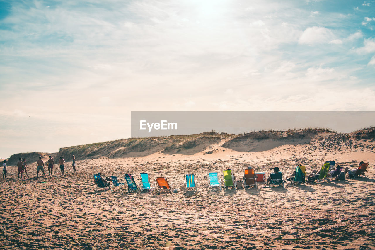 People relaxing on wooden chairs at beach against sky