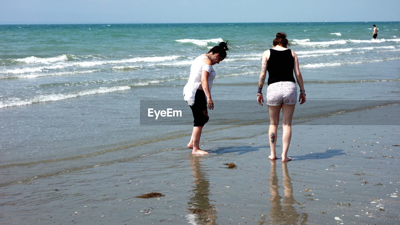 REAR VIEW OF WOMEN STANDING ON BEACH AGAINST SEA