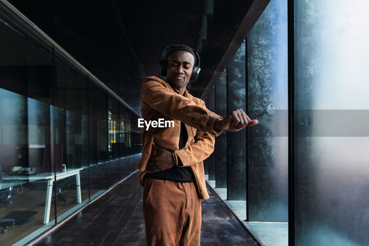 Stylish african american male dancing and listening to music in wireless headphones while standing near modern building with glass wall