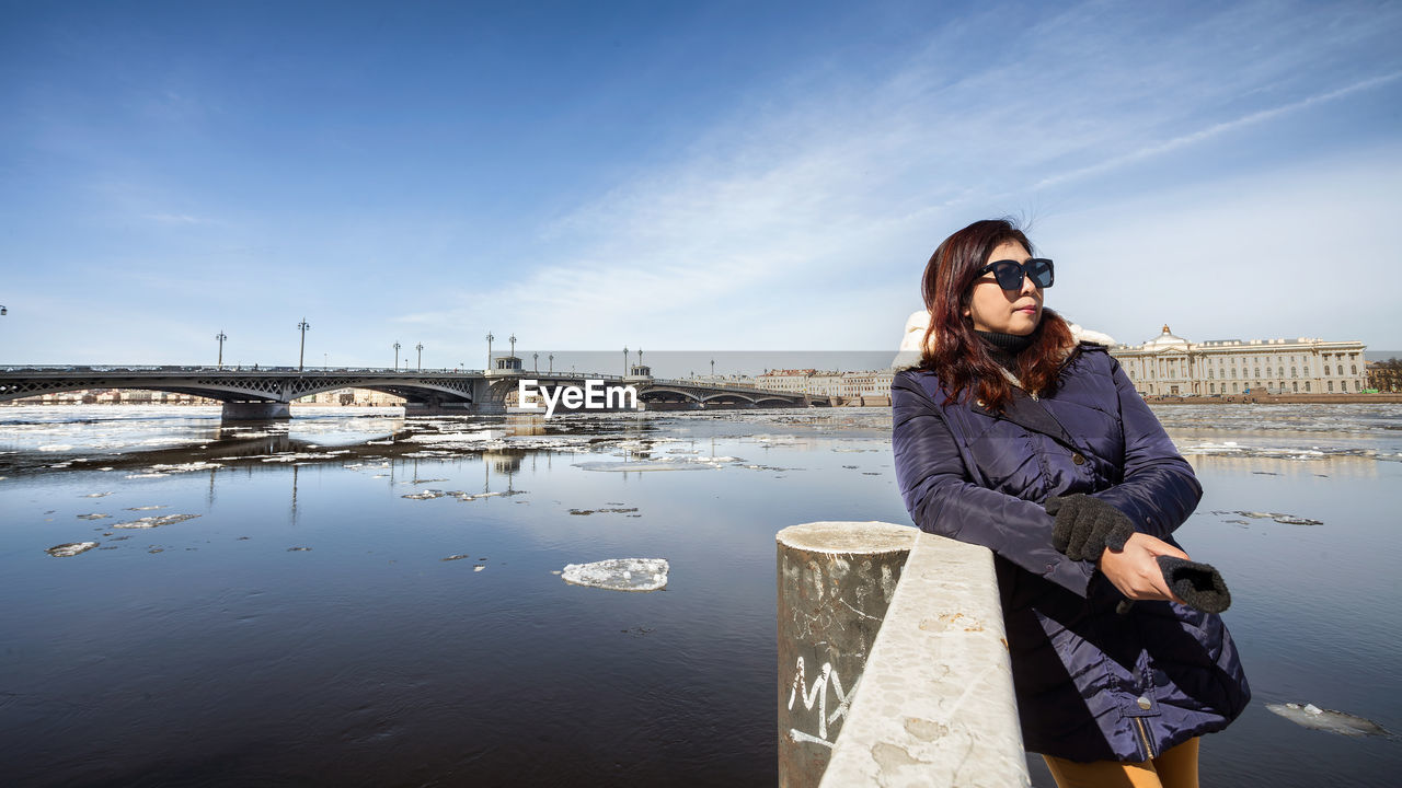 YOUNG WOMAN STANDING ON BRIDGE OVER WATER AGAINST SKY