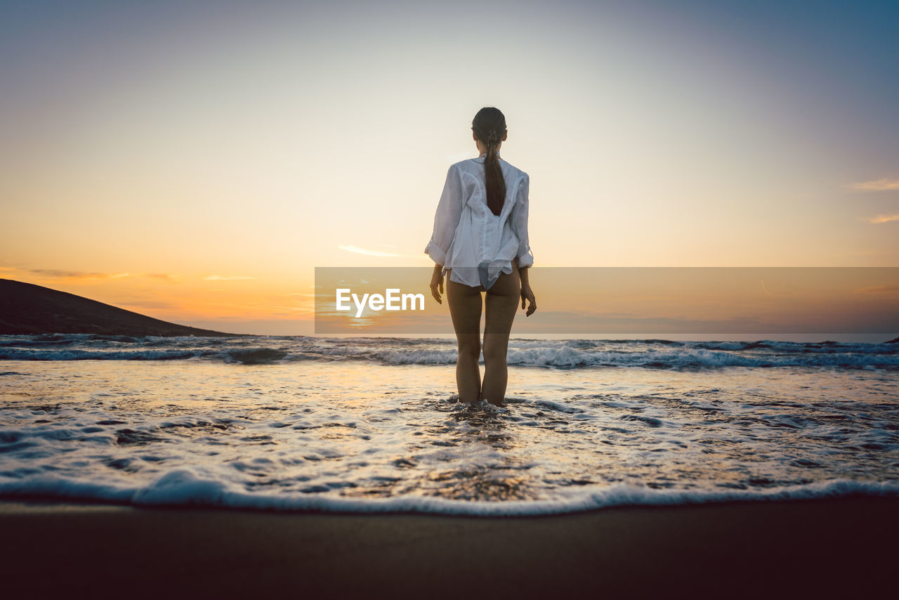 Rear view of seductive woman standing on shore at beach during sunset