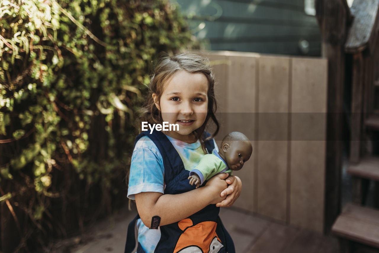 Little girl carrying babydoll in baby carrier outside in yard