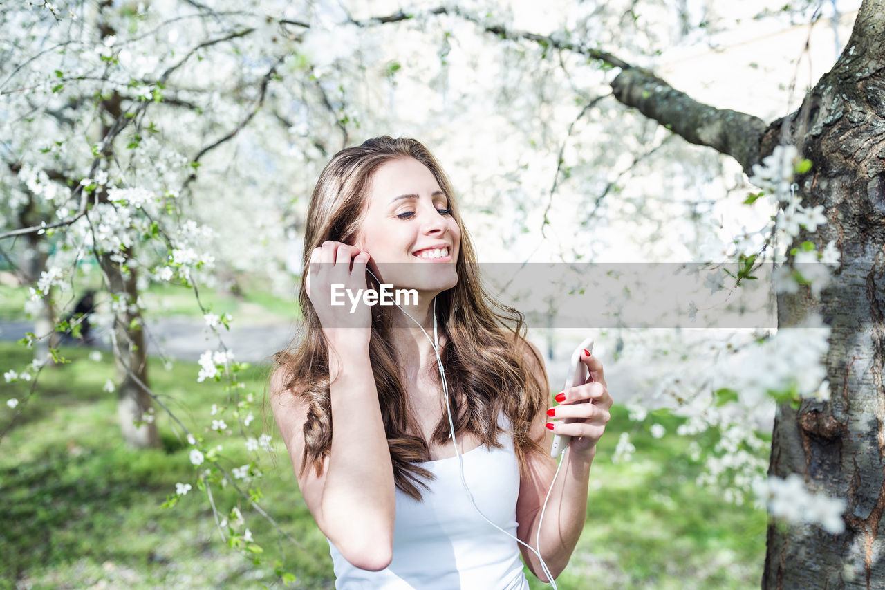 Young woman, closed eyes, holding phone, listening to music via earphones under the blooming trees