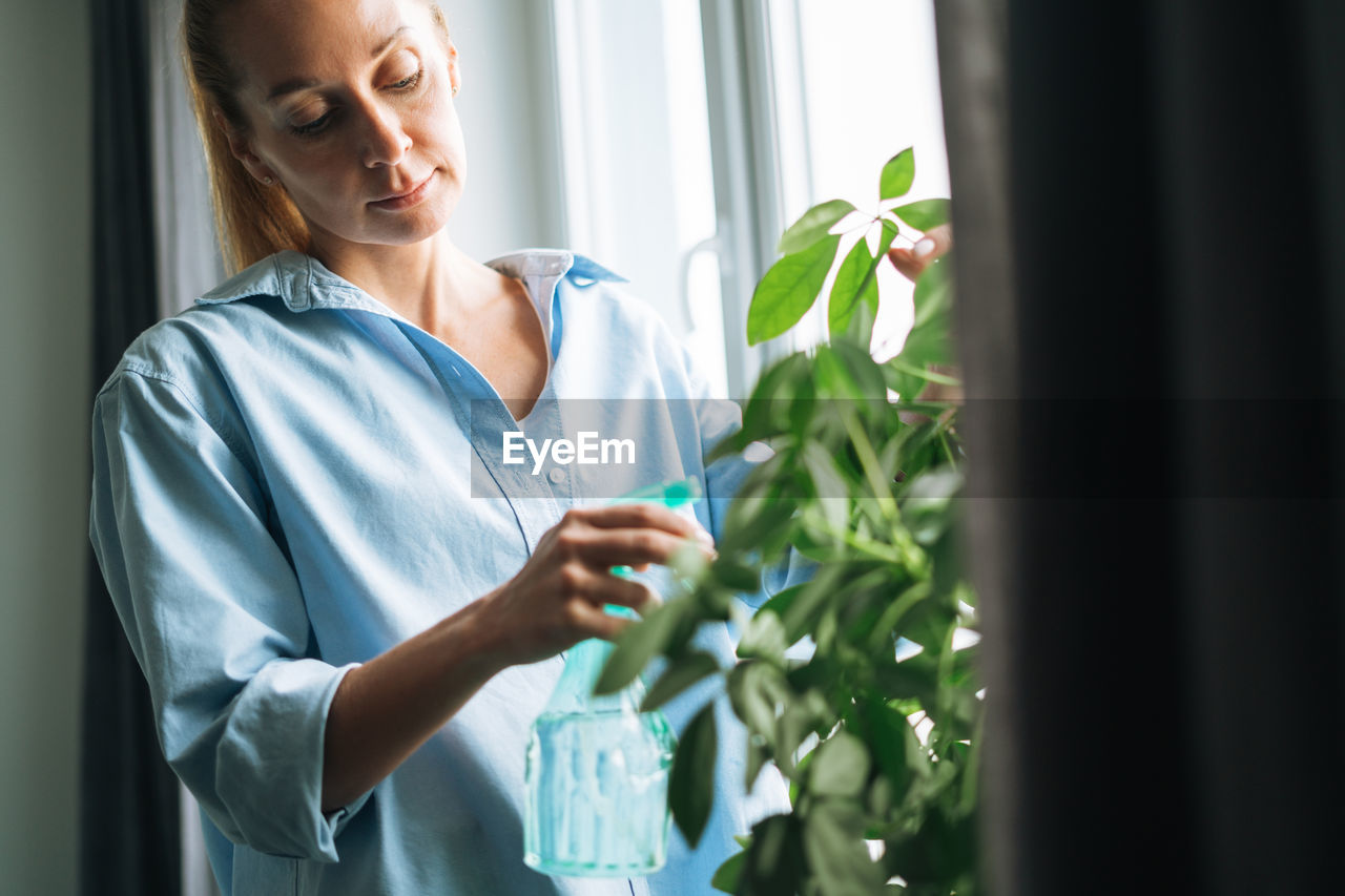 Young woman in blue shirt with spray with water takes care of houseplant in room at home