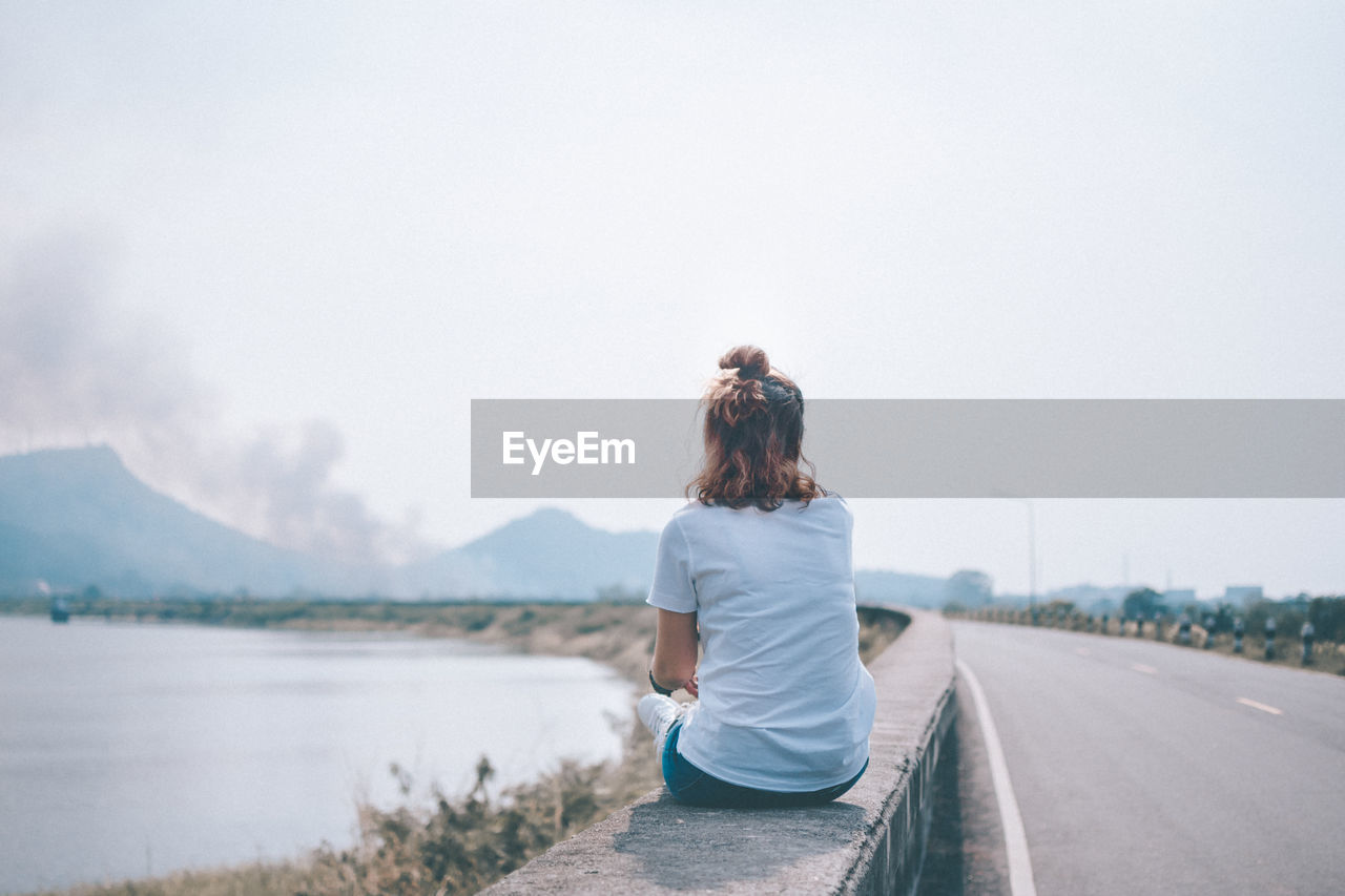 Rear view of woman sitting on retaining wall at lake against sky