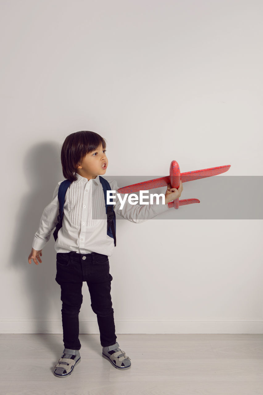 Boy child in a white shirt and jeans flies on a red airplane toy at the white wall of the house