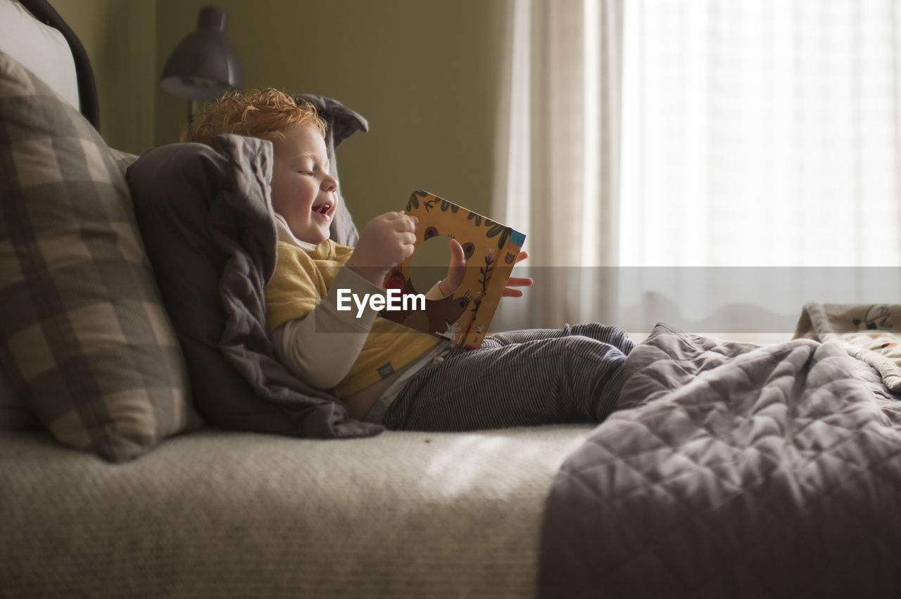 Little boy happily looks through picture book at home on his bed