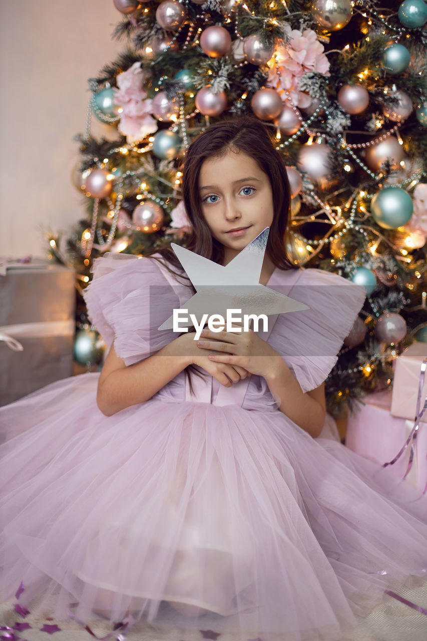 Girl child in a pink dress sits at the christmas tree and holds a star in her hands near her face