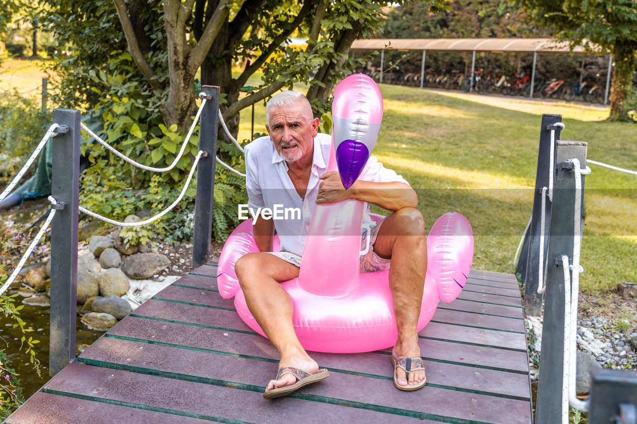 Portrait of smiling handsome middle aged man sitting on an inflatable pink flamingo toy