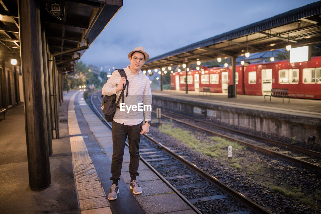 Portrait of mid adult man with backpack standing at railroad station platform against sky