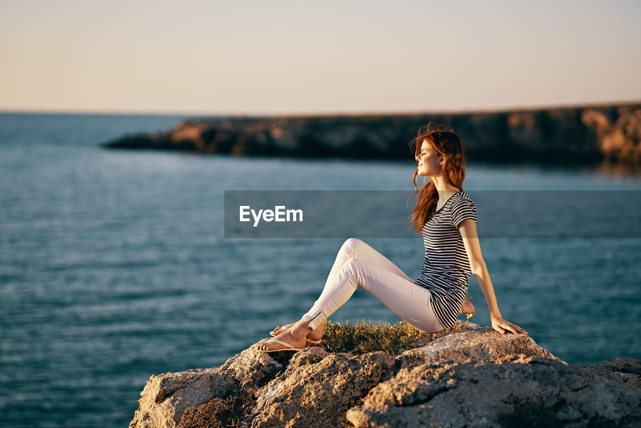 WOMAN SITTING ON ROCK AT SEA SHORE AGAINST SKY