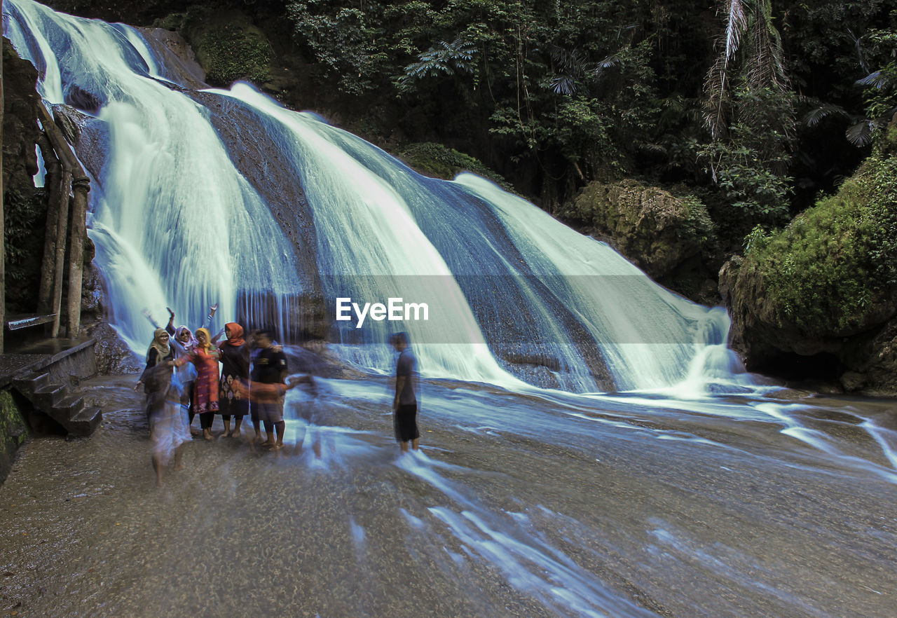 Long exposure of friends standing by waterfall in forest