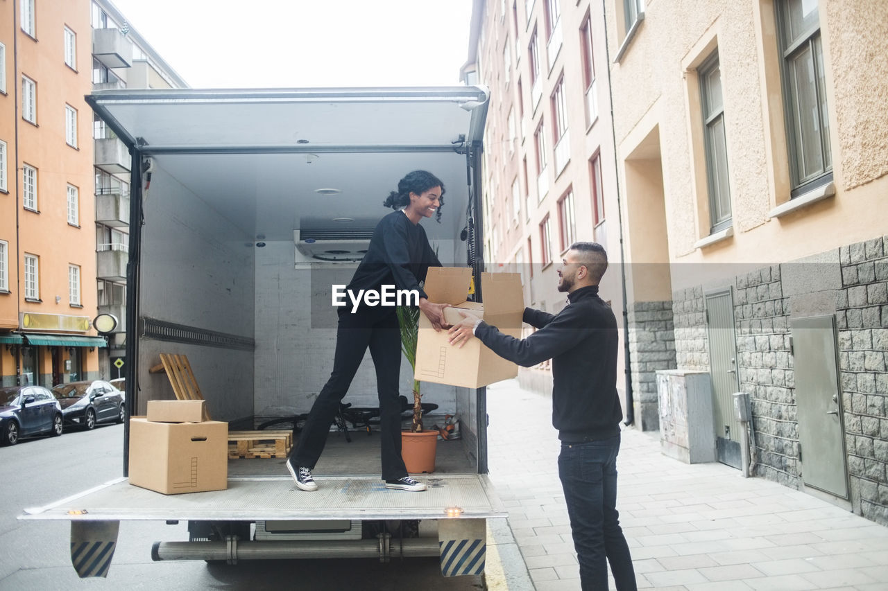 Smiling male and female movers unloading box from truck on street in city