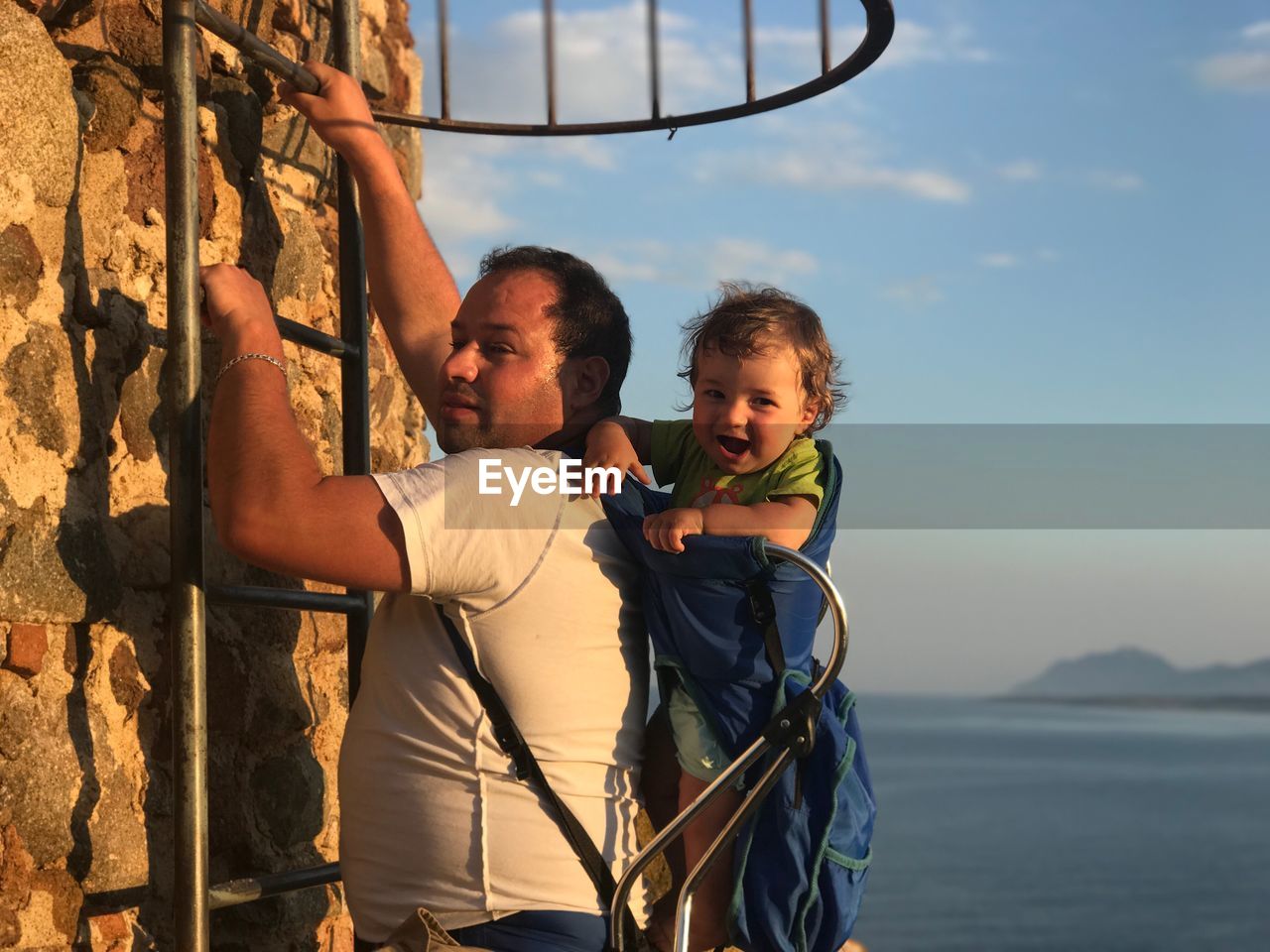 Father climbing ladder on wall while carrying son against sky