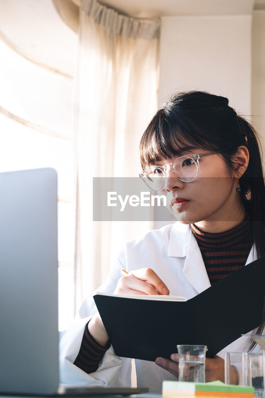 Scientist writing in book while looking at laptop at laboratory