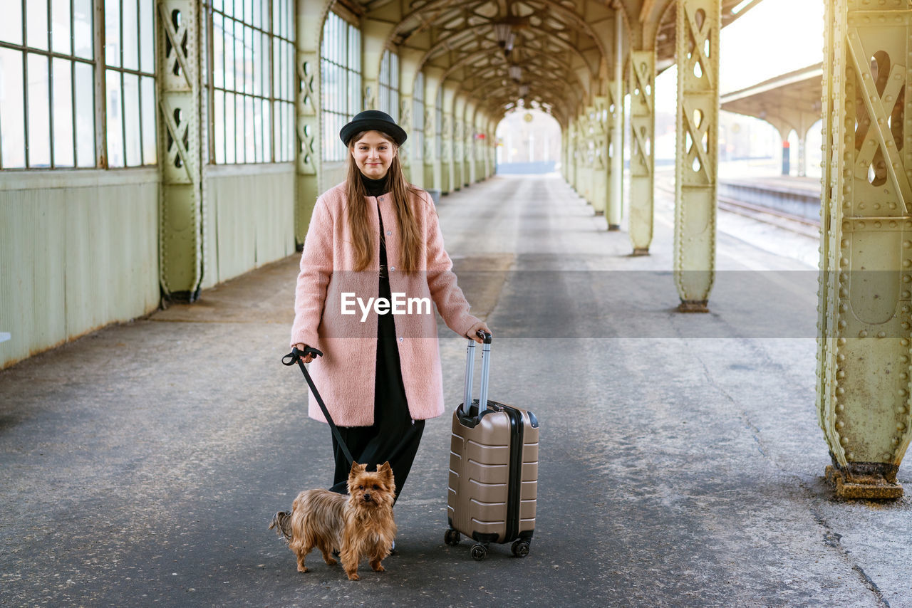 Beautiful young casual tourist woman with dog and suitcase waiting for train