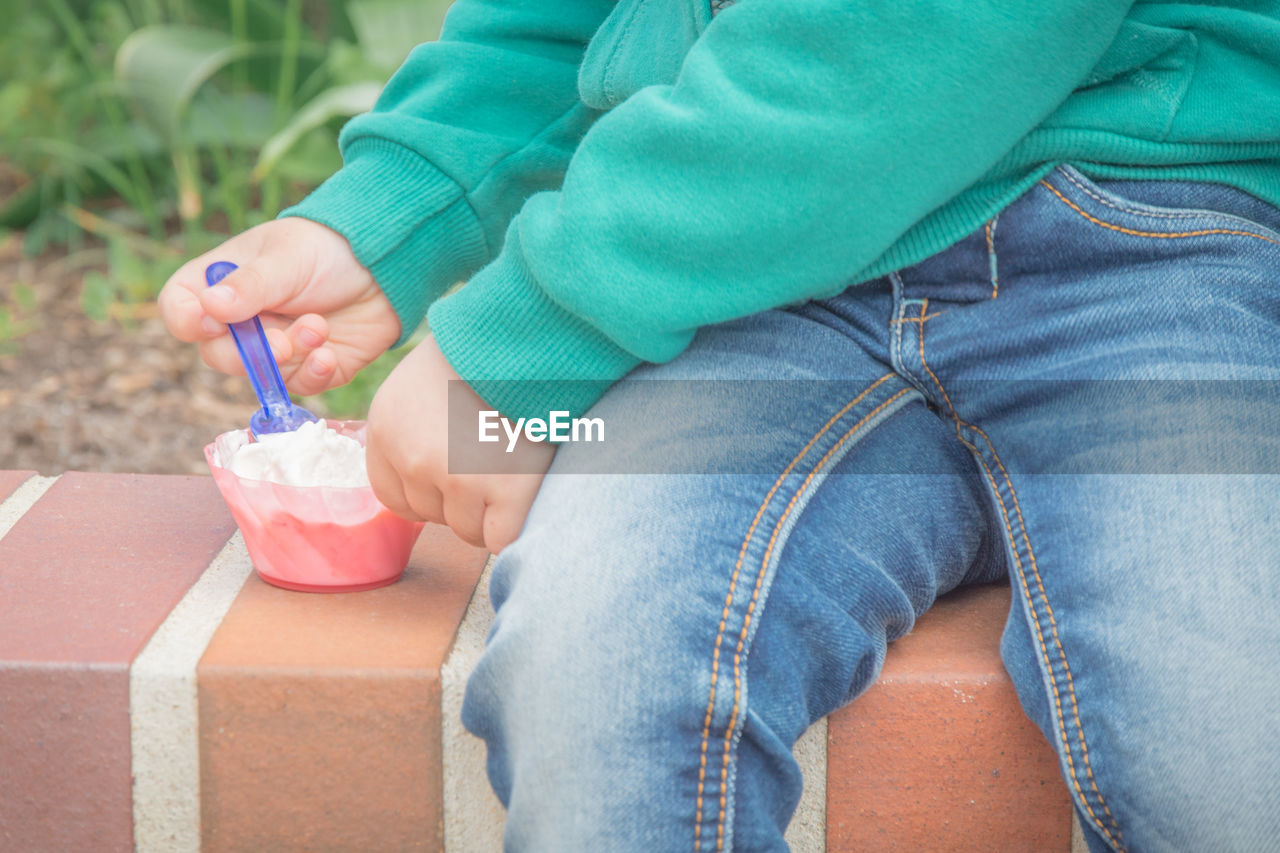 Midsection of person having ice cream in bowl while sitting on retaining wall