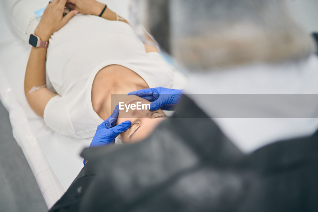 From above of beautician in gloves doing face massage for relaxed female client lying on table in beauty clinic