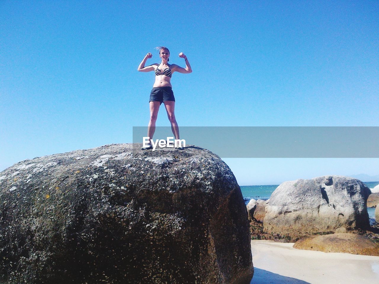 Low angle view of woman flexing muscles while standing on rock against sky 