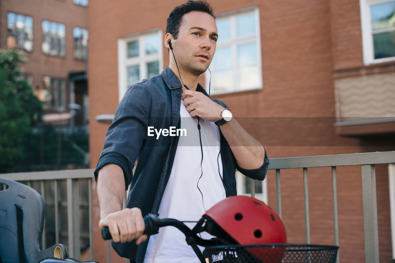 Mid adult man adjusting in-ear headphones while walking with bicycle against building in city