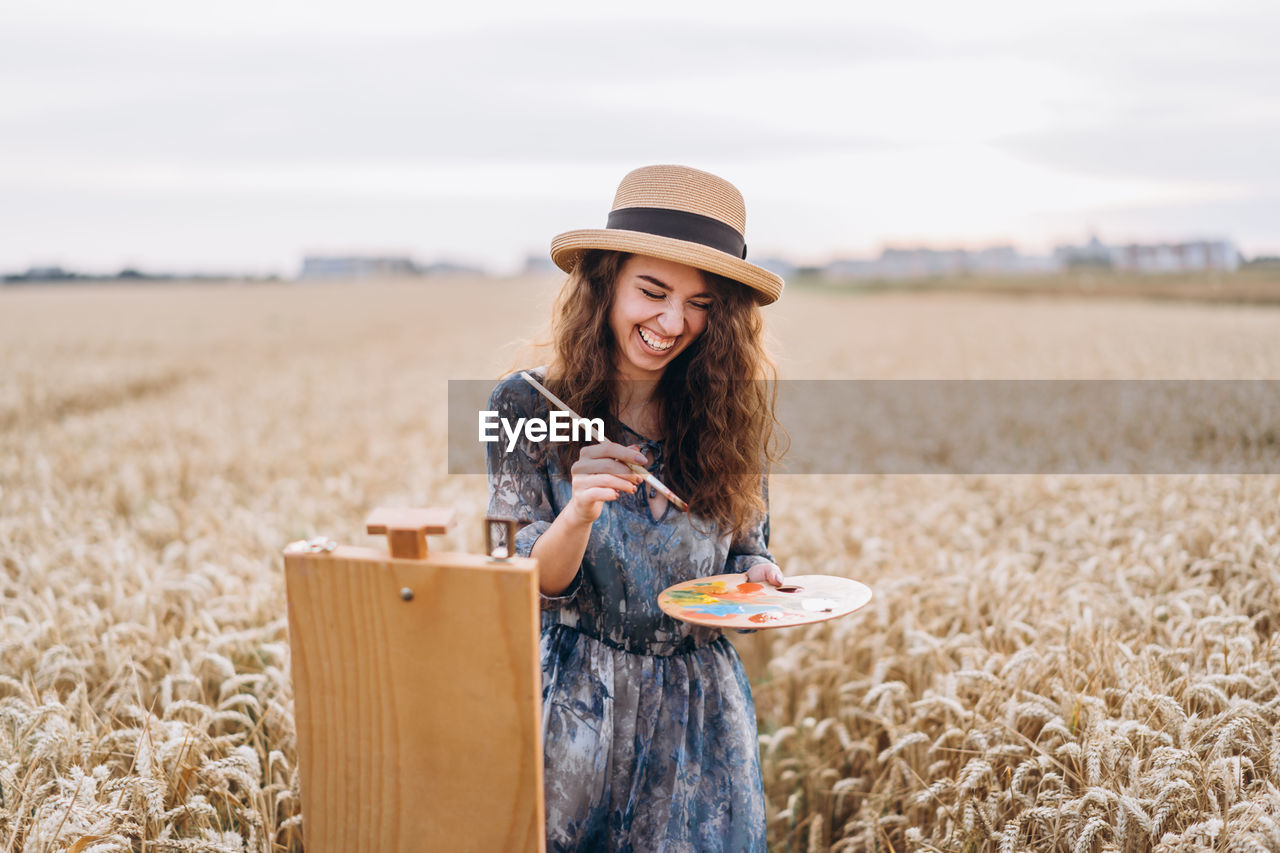 A young woman with curly hair and wearing a hat is painting in nature. 