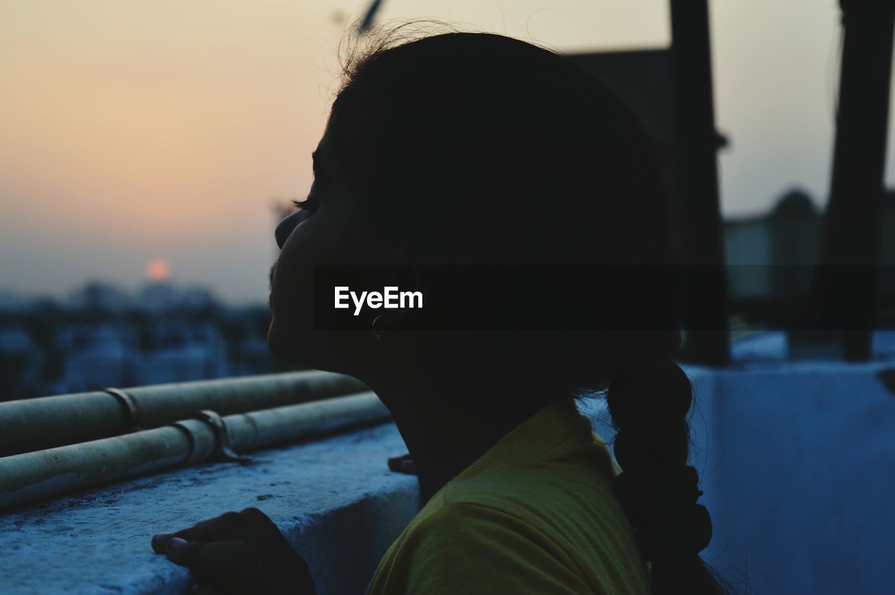 Side view of girl standing by retaining wall at building terrace during sunset