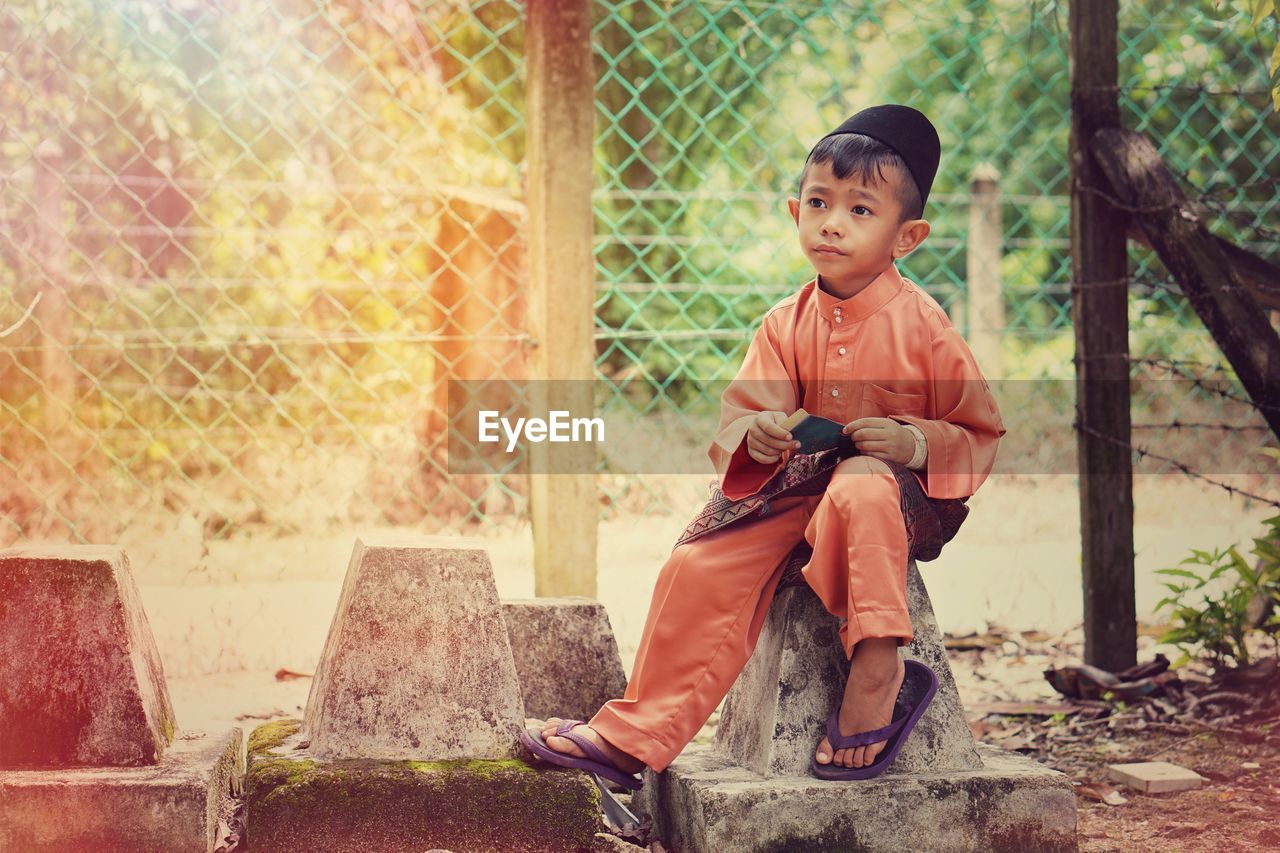 Thoughtful boy sitting on stone against fence