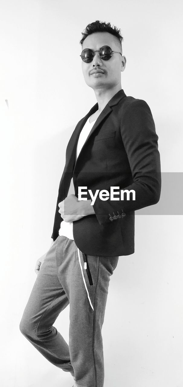 PORTRAIT OF YOUNG MAN WEARING SUNGLASSES STANDING AGAINST WALL