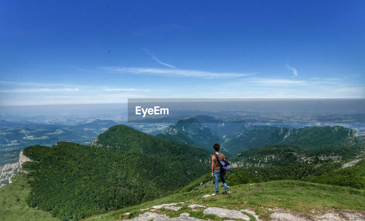 REAR VIEW OF MAN STANDING ON MOUNTAINS AGAINST SKY