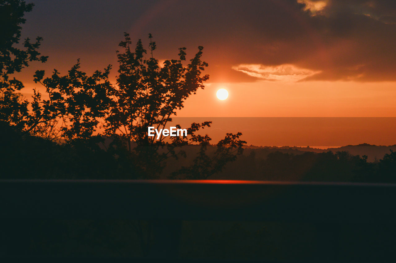 Silhouette trees by lake against orange sky