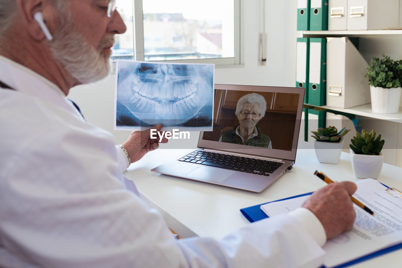Side view of elderly male doctor in wireless earbud speaking with woman on video call on netbook in clinic