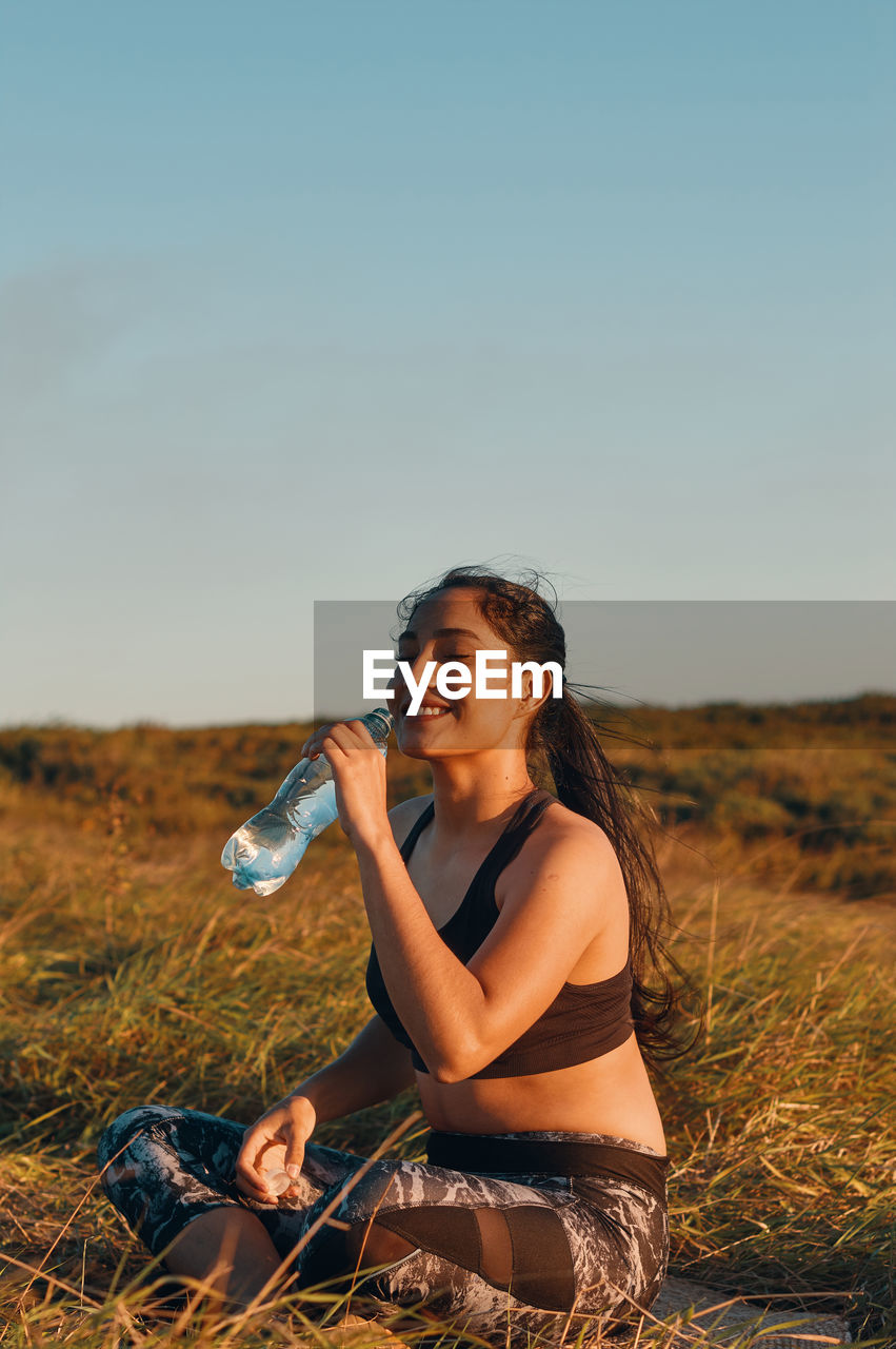 Smiling woman drinking water while sitting on grass against sky