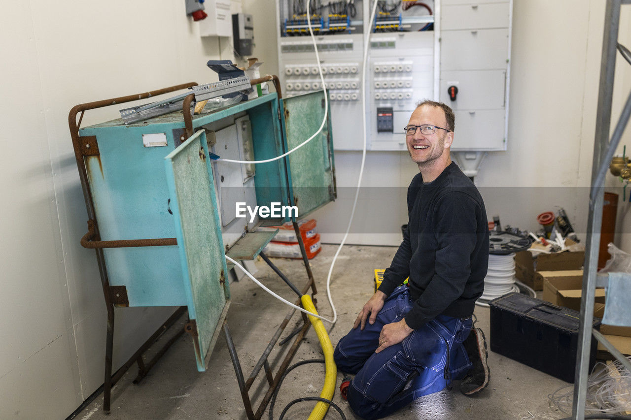 Smiling male electrician kneeling by electric panel in industry