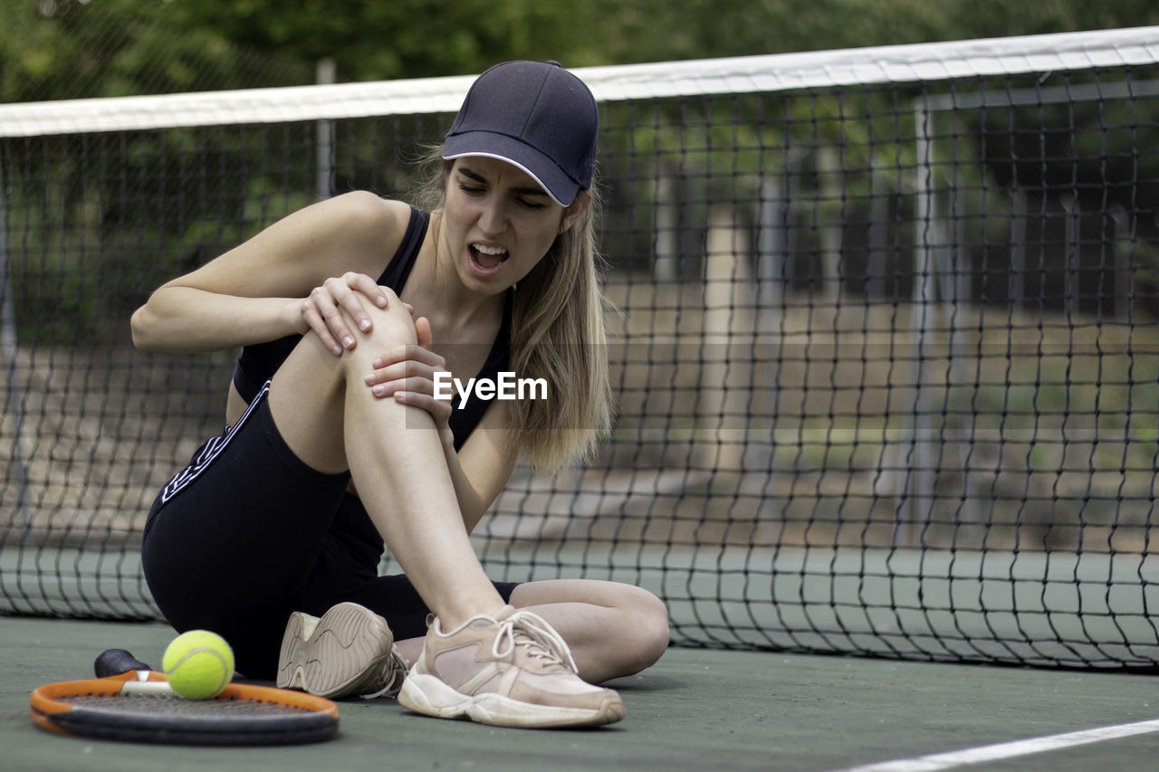 Young female tennis player sitting on court injured knee and showing a face of pain