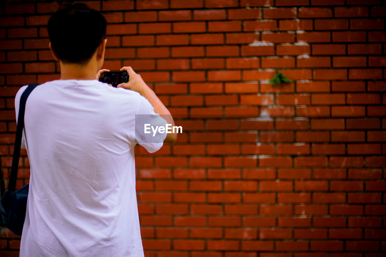 Rear view of man photographing against brick wall