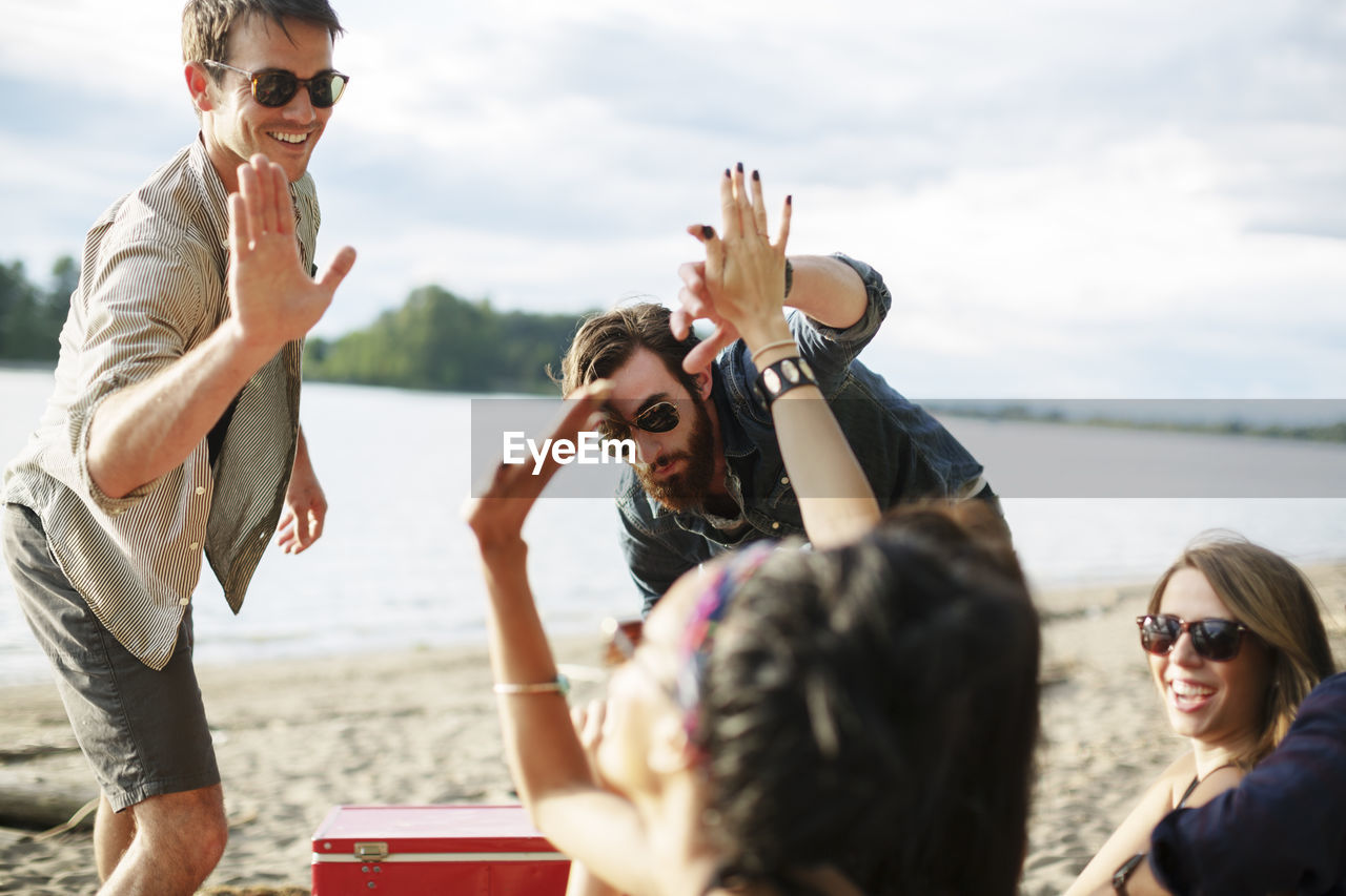 Happy friends doing high-five while enjoying at riverbank against cloudy sky