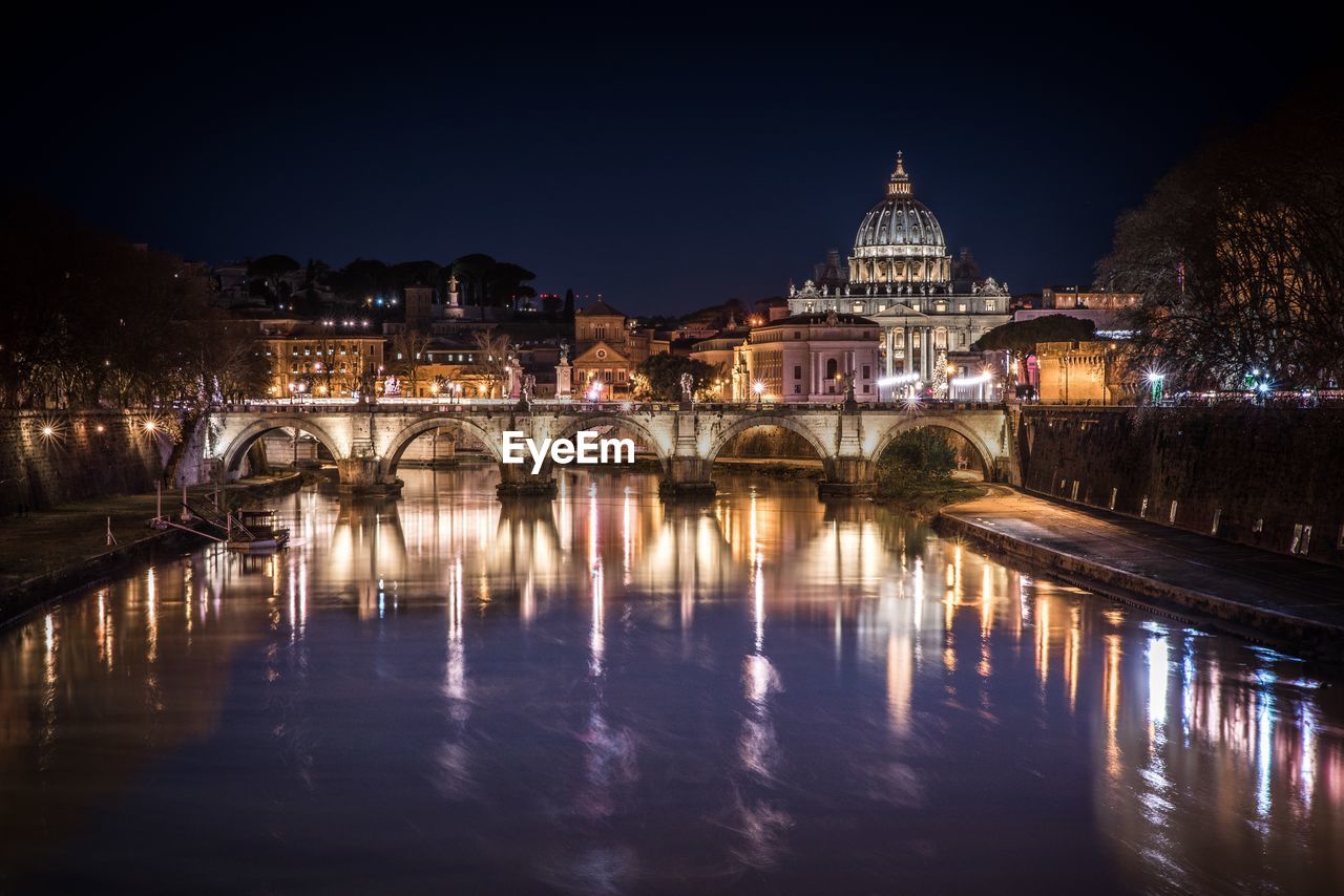 Illuminated bridge over river by buildings against sky at night