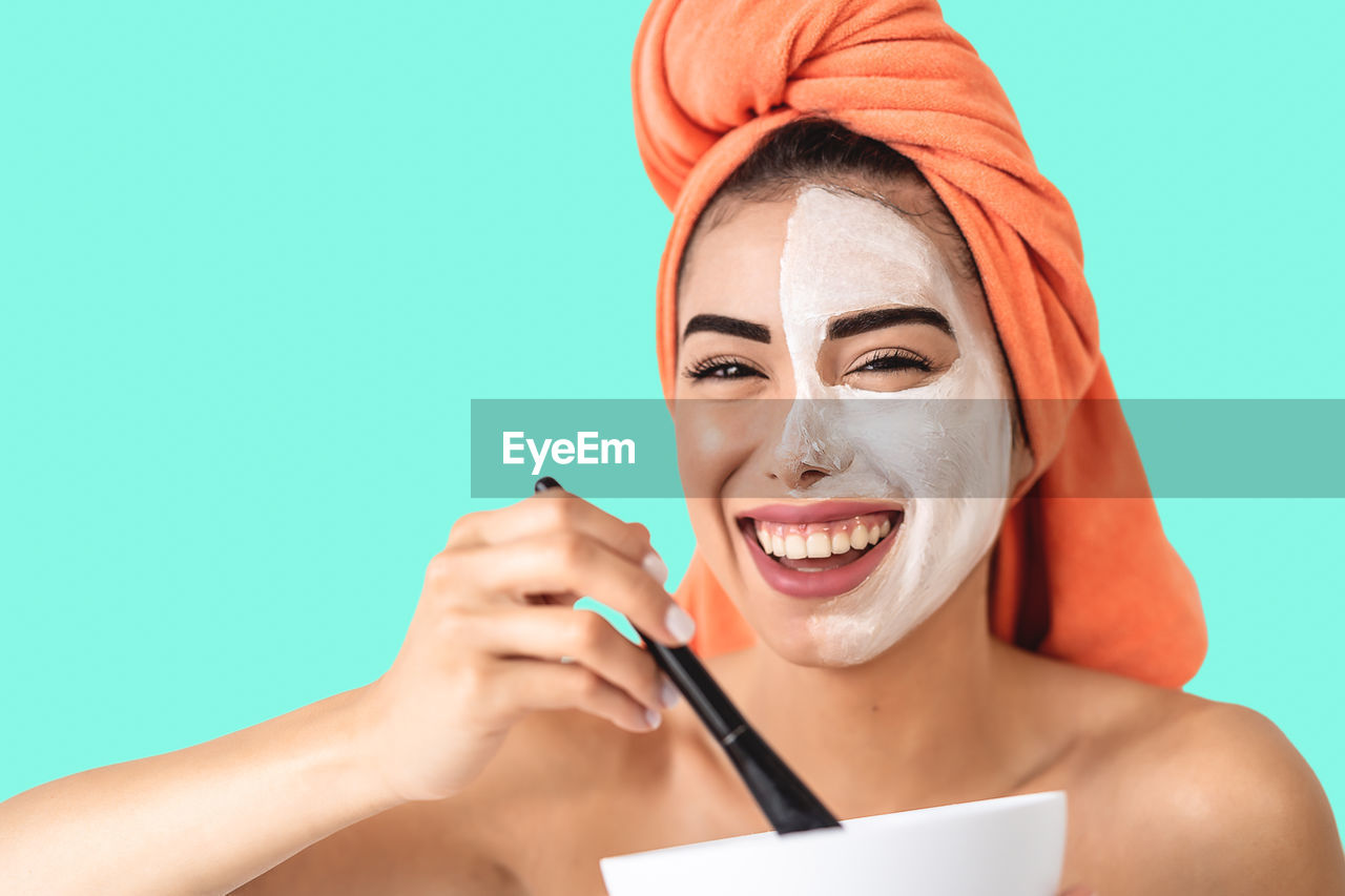 Portrait of happy young woman applying facial mask against blue background