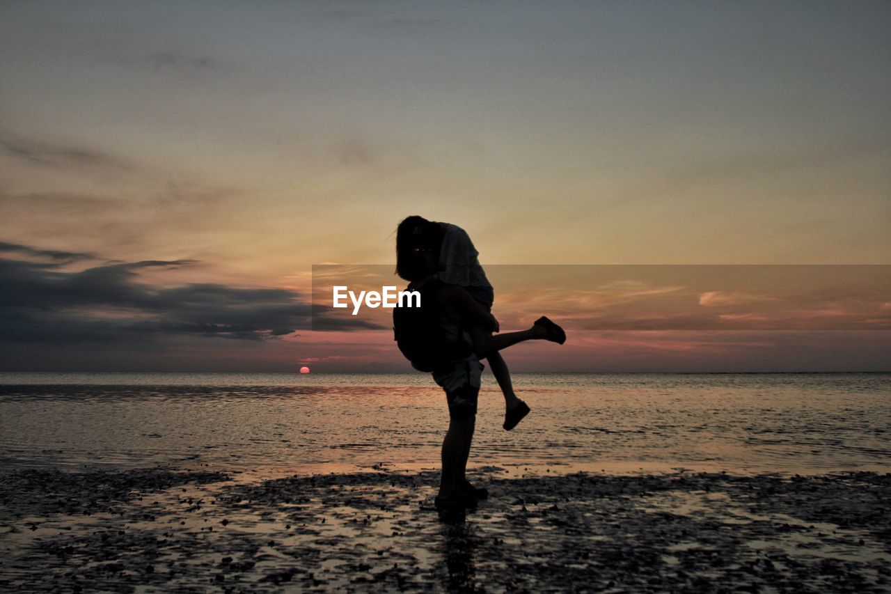 Man lifting woman on wet shore at beach against sky during sunset