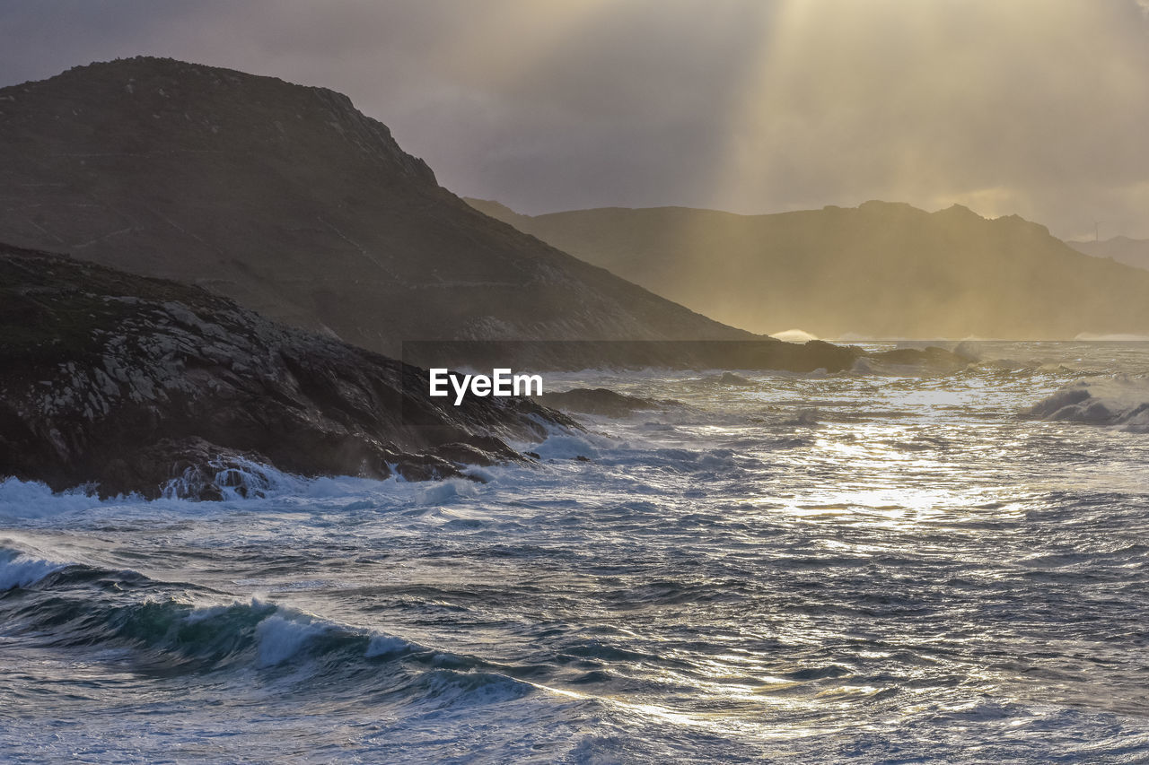 Big waves break on the coast, in the sunlit sea, passing through the clouds sunset. galicia, spain.