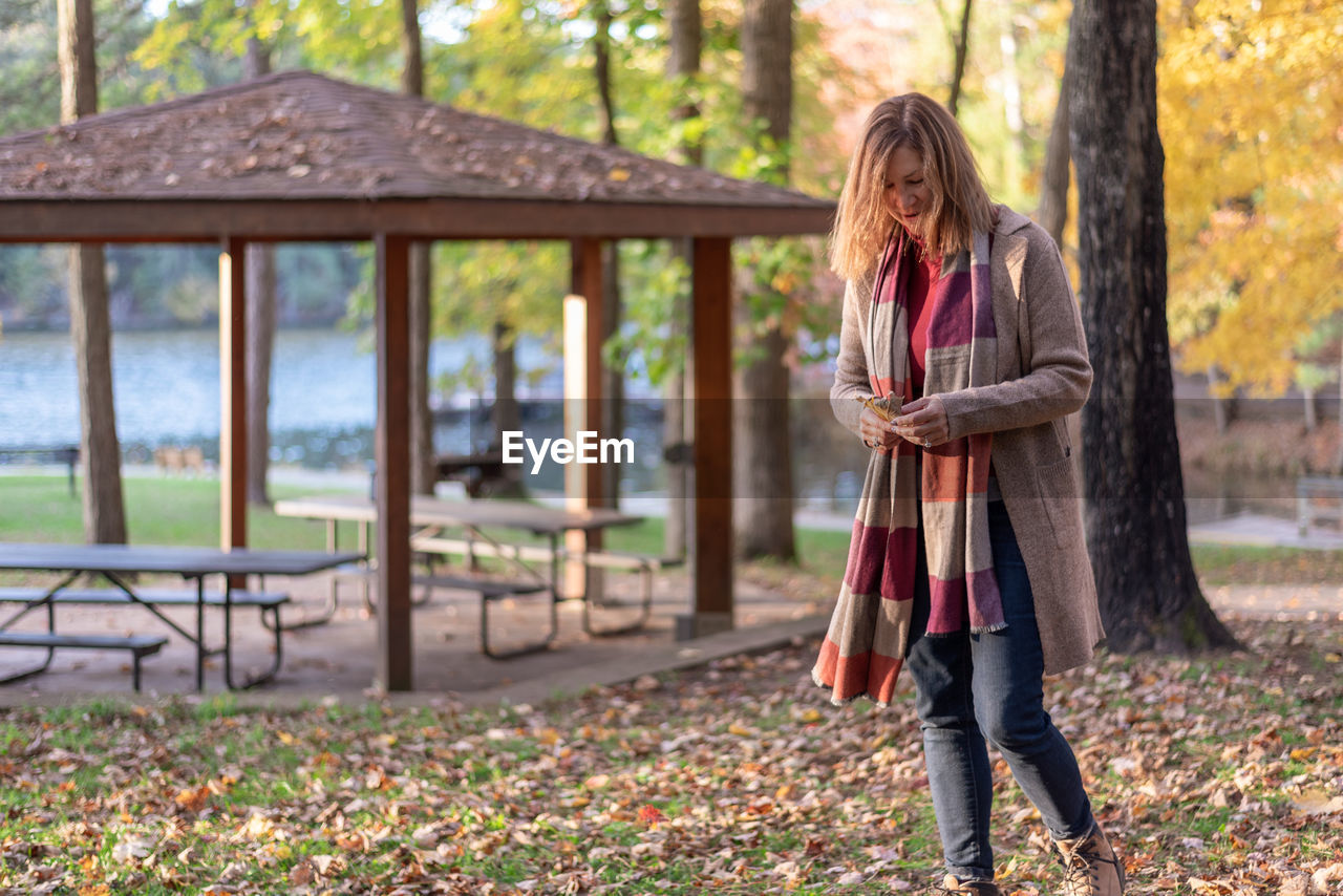 Full length of mature woman standing in park while holding maple leaf during autumn