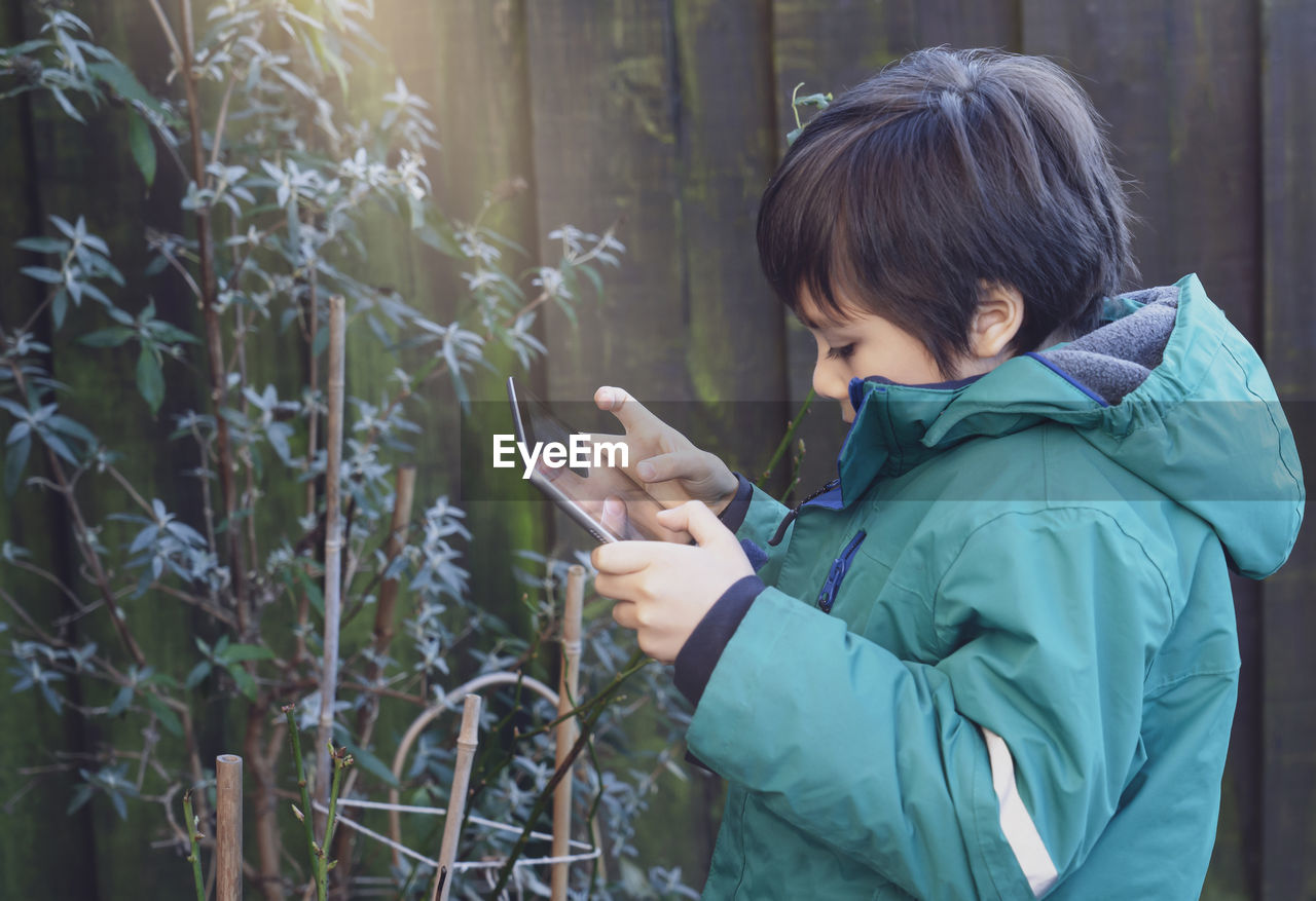 Side view of boy using digital tablet by plants outdoors