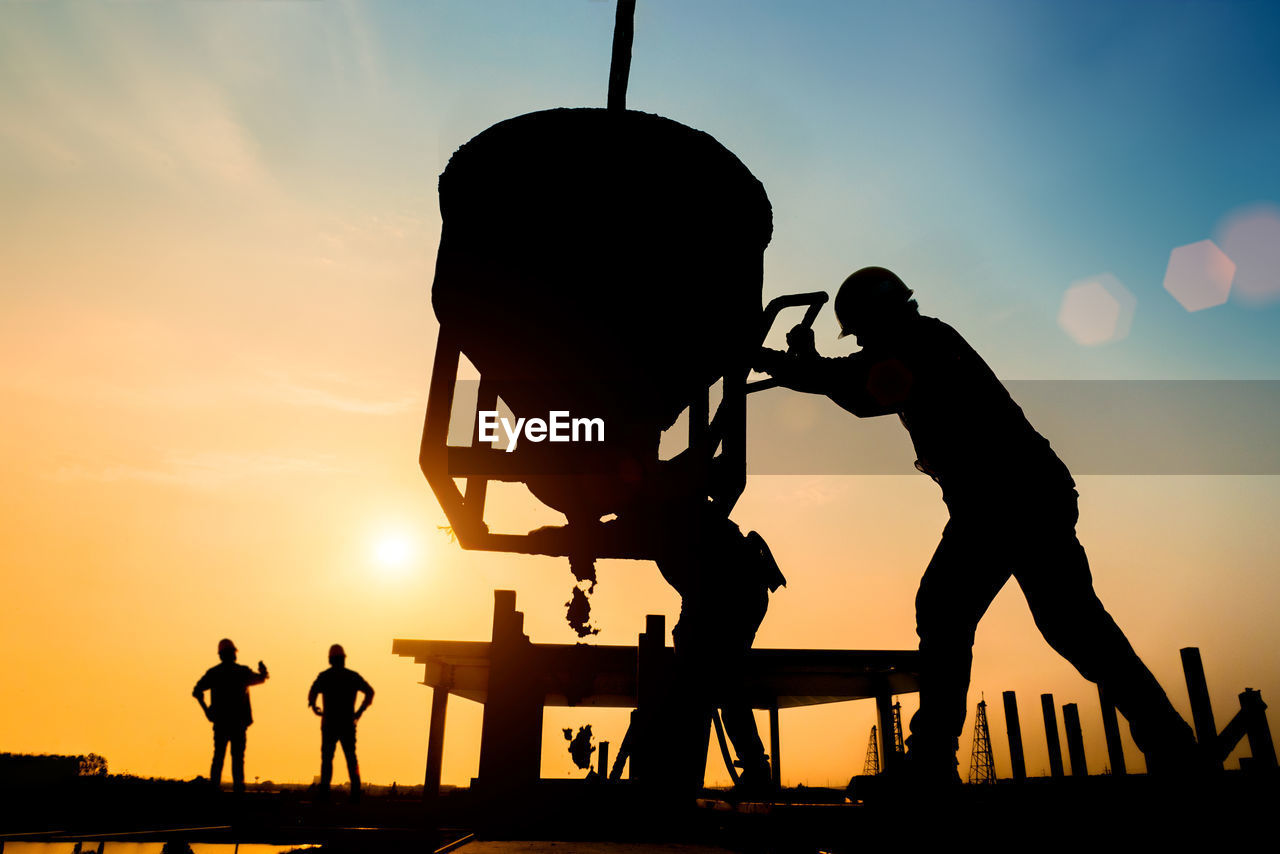 Silhouette construction worker pouring concrete on site against sky during sunset