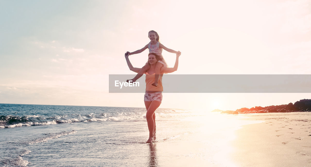 Full length of woman carrying happy daughter on shoulder at beach