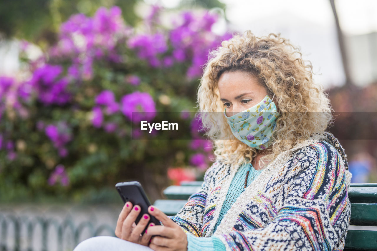 Smiling woman wearing mask using mobile phone while sitting outdoors