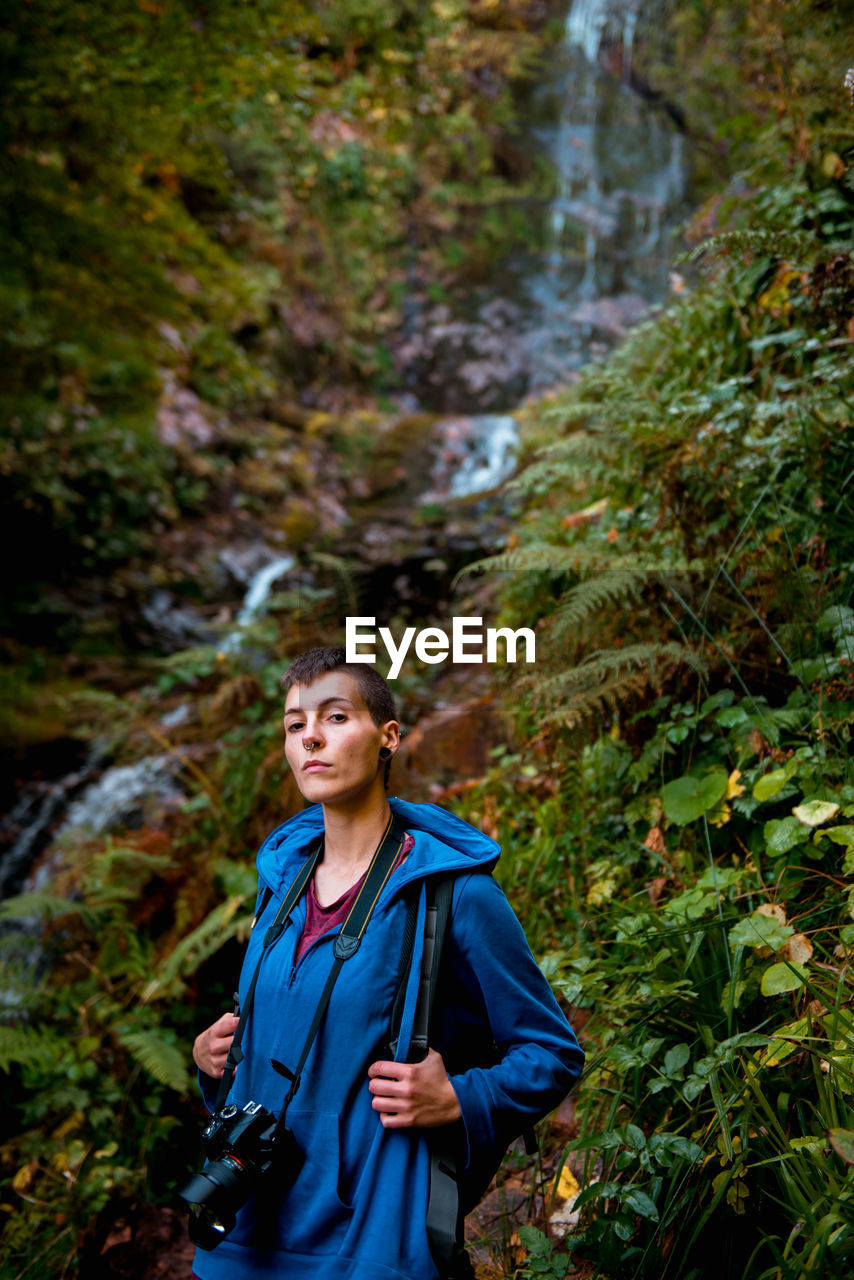 Focused young woman with professional camera and backpack looking at camera while standing at waterfall in forest in summer day