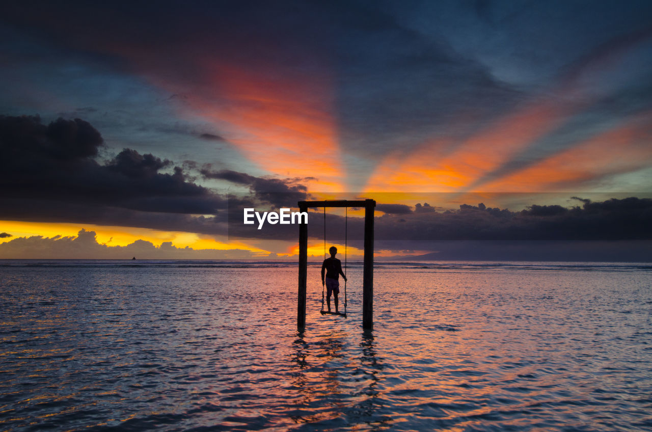 Silhouette man swinging over sea against cloudy sky during sunset