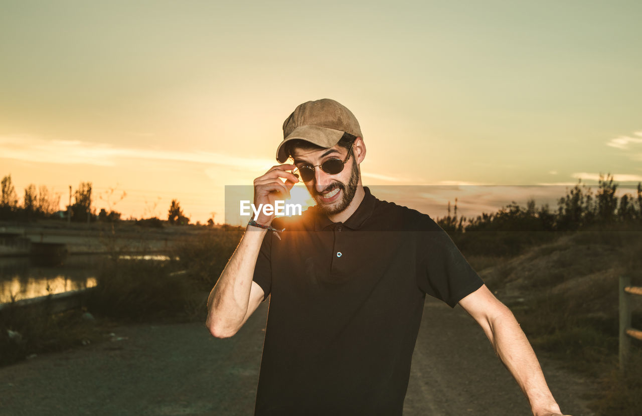 Portrait of young man wearing sunglasses while standing against sky during sunset